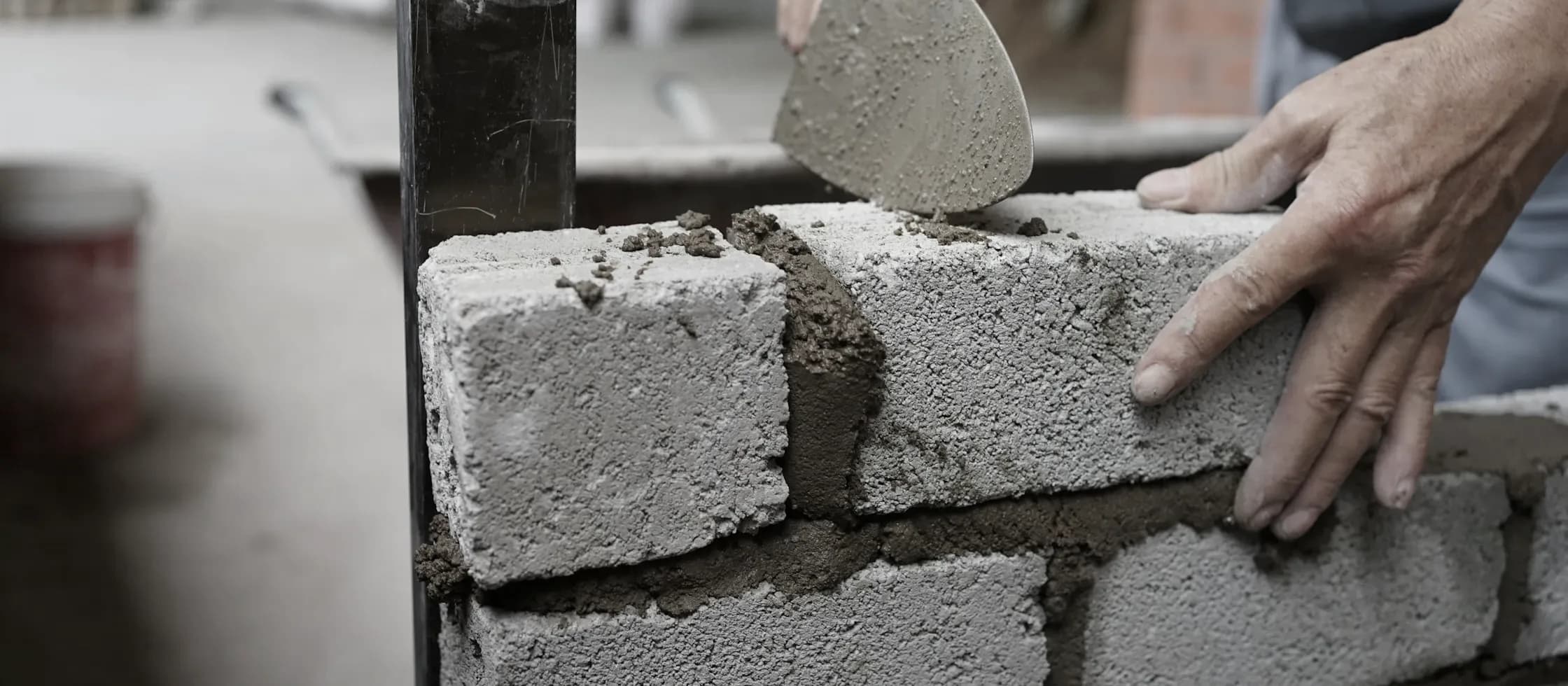 Hands bedding a concrete block onto a mortar course, a trowel held above the joint.