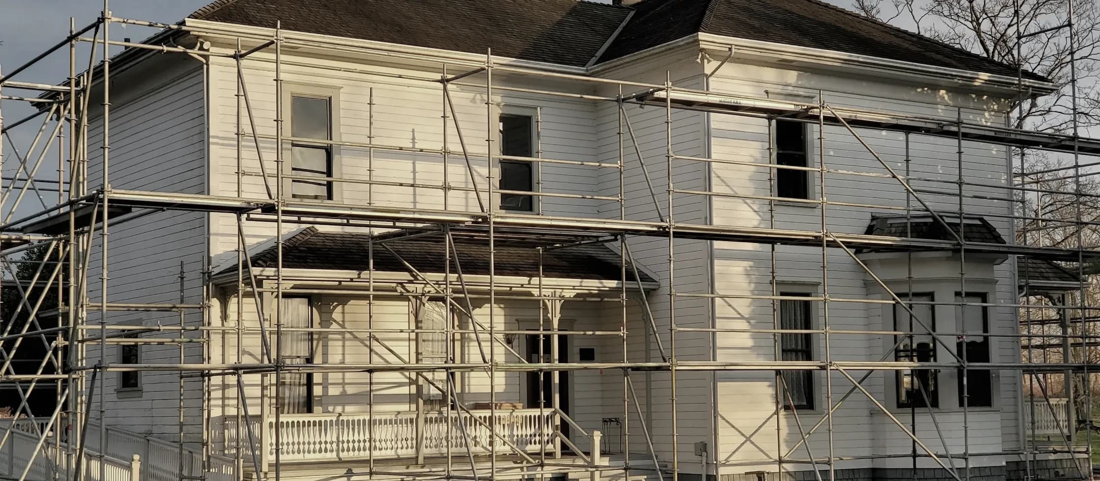 A clapboard house wrapped in tube scaffolding in late afternoon light, mid-renovation.